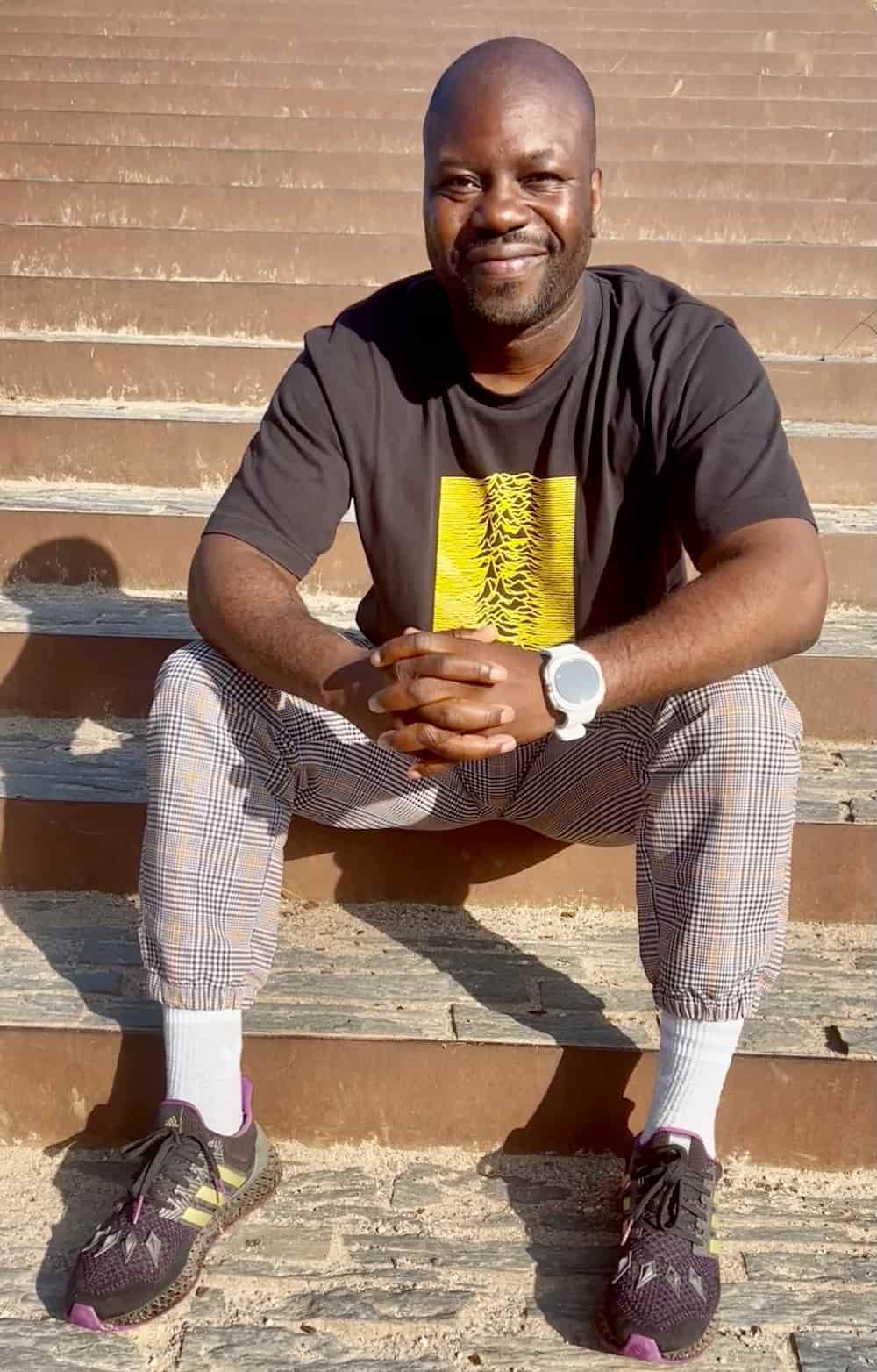 FIGパルクール委員長のシャルル・ペリエール氏が階段に腰掛けている写真。黒いTシャツに黄色のデザインが入り、チェック柄のパンツとスニーカーを着用。FIGアカデミーやパルクール教育プログラムの確立に尽力し、パルクールを次世代へと安全に継承することに取り組んでいる重要人物。日なたの中でリラックスした表情を見せている。